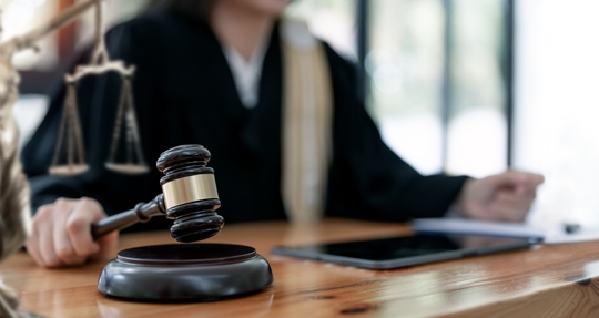 A wooden gavel and sound block are in the foreground, with a judge in a robe and a scale of justice in the blurred background. | Mathur Law Offices, P.C.