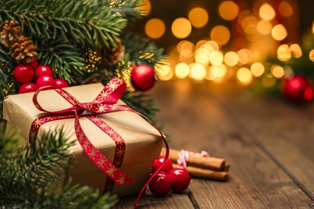 A wrapped Christmas gift with a red ribbon sits on a wooden surface surrounded by pine branches, red ornaments, and warm holiday lights in the background. | Mathur Law Offices, P.C.